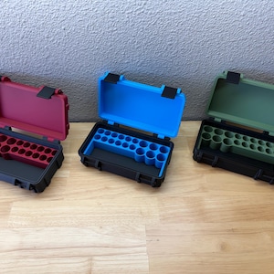 May include: Three rectangular storage cases with black outer shells and hinged lids. Each case features a colored interior insert with multiple circular cutouts for organization. The inserts are available in matte red, bright blue, and olive green.