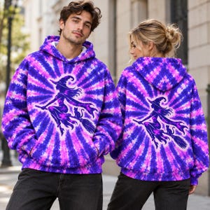 Könnte beinhalten: Ein lila und pinkfarbener Batik-Kapuzenpullover mit einer Grafik einer Hexe auf einem Besen mit einer Katze. Das Design strahlt in einem Sonnenstrahlenmuster von der Mitte aus. Der Kapuzenpullover hat eine Kapuze mit Kordelzug und eine Kängurutasche.