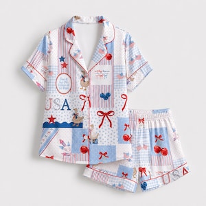 Op de afbeelding: Een pyjamaset met korte mouwen en knoopsluiting, voorzien van een patriottische print met ganzen, rode strikken, kersen en Amerikaanse vlaggen. De witte stof bevat blauwe en rode strepen, sterren en de tekst USA en God Bless America, Land of the Free.