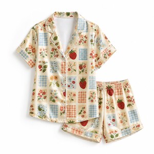 May include: A short-sleeve button-down pajama shirt and matching elastic-waist shorts set in a cream color. The fabric features a repeating grid pattern of strawberries, various small colorful flowers, and blue or orange checkered squares.