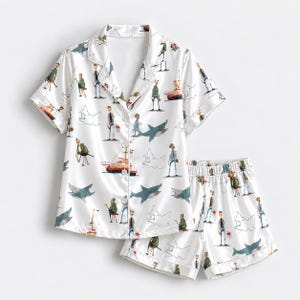 May include: A white short-sleeved button-down pyjama shirt and matching shorts set featuring a repeating pattern of illustrated figures, fishing boats, and sharks in shades of blue, red, and green. The shirt has a notched collar and white buttons.