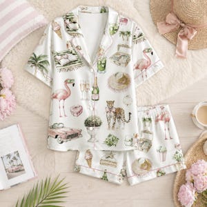 Puede incluir: Un conjunto de pijama blanco de manga corta con botones y pantalón corto a juego, con un estampado repetitivo de flamencos rosas, un leopardo, un coche rosa vintage, sombreros de paja, palmeras, helados, botellas de champán y maletas.