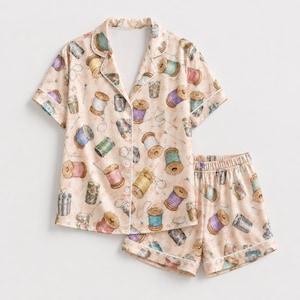 May include: A short-sleeve button-down pyjama shirt and matching elastic-waist shorts set in a light peach colour. The fabric features a repeating pattern of colourful sewing thread spools, thimbles, and needles scattered across the entire garment.