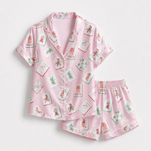 Pode incluir: Um conjunto de pijama rosa claro composto por uma camisa de manga curta com botões e calções a condizer. O tecido apresenta uma estampa repetida de cartas de jogar, peças de mahjong com a palavra NORTH e ilustrações natalícias em vermelho e verde.