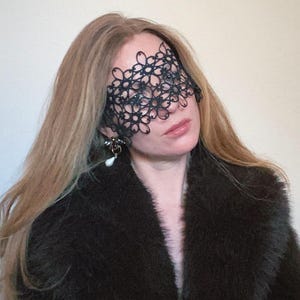 May include: A woman wearing a black fur coat and a black lace eye mask. The mask covers her eyes and part of her forehead.
