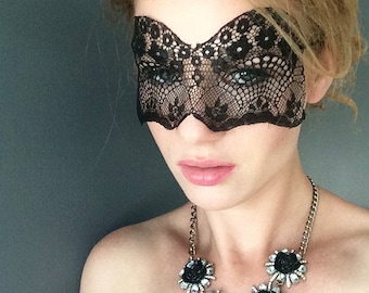 May include: A woman wearing a black lace eye mask and a gold necklace with black and clear rhinestone flower accents.