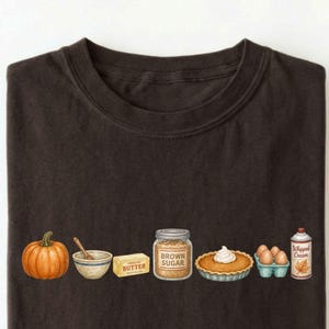 May include: A dark brown t-shirt featuring a horizontal graphic of baking ingredients including an orange pumpkin, a mixing bowl with a spoon, a stick of butter, a jar labeled Brown Sugar, a pumpkin pie, a carton of eggs, and whipped cream.