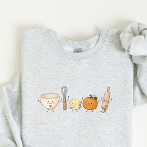 Puede incluir: Una sudadera gris claro de cuello redondo con un estampado gráfico de cinco utensilios de cocina antropomórficos: un bol, un batidor, un trozo de mantequilla, una calabaza y un rodillo, todos con caras sonrientes y pequeños corazones dibujados.