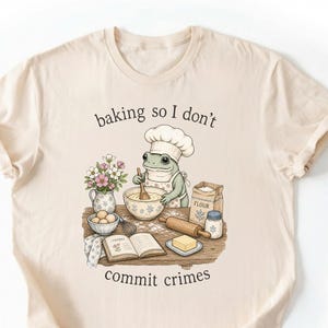 Könnte beinhalten: Ein cremefarbenes T-Shirt mit einer Grafik eines Frosches, der eine Kochmütze und eine Schürze trägt und an einem Holztisch mit einer Schüssel, Mehl, Eiern und einem Rezeptbuch backt. Der Text lautet, baking so I don't commit crimes.