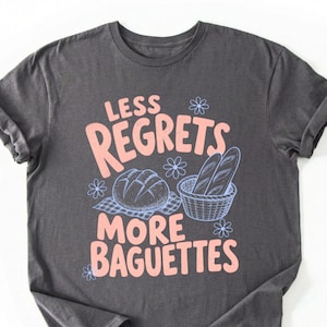 May include: A charcoal gray short-sleeve t-shirt featuring a graphic print in light pink and light blue. The text reads LESS REGRETS MORE BAGUETTES, accompanied by illustrations of a round loaf of bread and a basket of baguettes with small flowers.