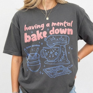 May include: A charcoal gray short-sleeve t-shirt featuring a graphic print in pink and light blue. The text reads having a mental bake down and from scratch. The design includes a stand mixer, stacked bowls, whisks, and a cookie cooling rack.