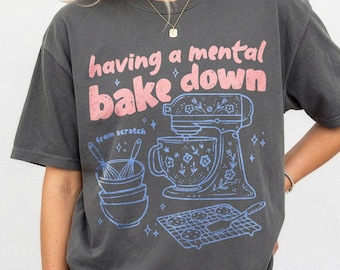 May include: A charcoal gray short-sleeve t-shirt featuring a graphic print in pink and light blue. The text reads having a mental bake down and from scratch. The design includes a stand mixer, stacked bowls, whisks, and a cookie cooling rack.