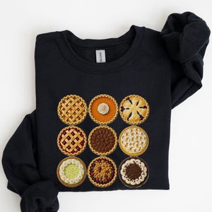 Puede incluir: Una sudadera negra de cuello redondo con un estampado gráfico de nueve pasteles diferentes dispuestos en una cuadrícula de tres por tres. Los pasteles incluyen varios estilos de corteza, rellenos y coberturas como rejilla.