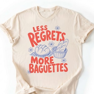 May include: A cream-coloured t-shirt featuring a graphic print with the red text "LESS REGRETS MORE BAGUETTES" and blue line-art illustrations of a round loaf of bread on a checkered cloth and two baguettes in a basket, accented by small blue flowers.