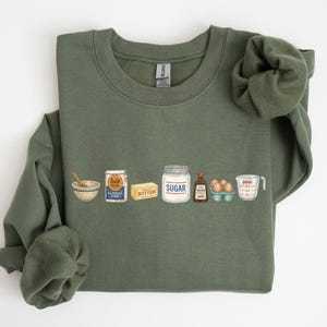 Könnte beinhalten: Ein olivgrünes Sweatshirt mit Rundhalsausschnitt und einem Grafikdruck von Backzutaten, darunter eine Rührschüssel mit Löffel, eine Packung Gold Medal Mehl, ein Stück Butter, ein Glas mit der Aufschrift Sugar, Vanilleextrakt, Eier und ein Messbecher.