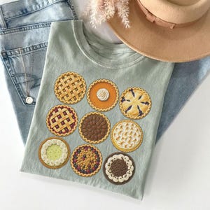 Puede incluir: Una camiseta verde salvia con un estampado gráfico de nueve pasteles diferentes, incluyendo tartas de frutas con corteza de rejilla, una tarta de calabaza con nata montada y tartas de chocolate, sobre unos vaqueros azul claro y un sombrero de fieltro beige.