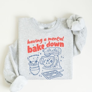 May include: A light gray crewneck sweatshirt featuring a red and blue graphic print. The text reads having a mental bake down and from scratch. The illustration includes a stand mixer, stacked mixing bowls with whisks, and a cooling rack.