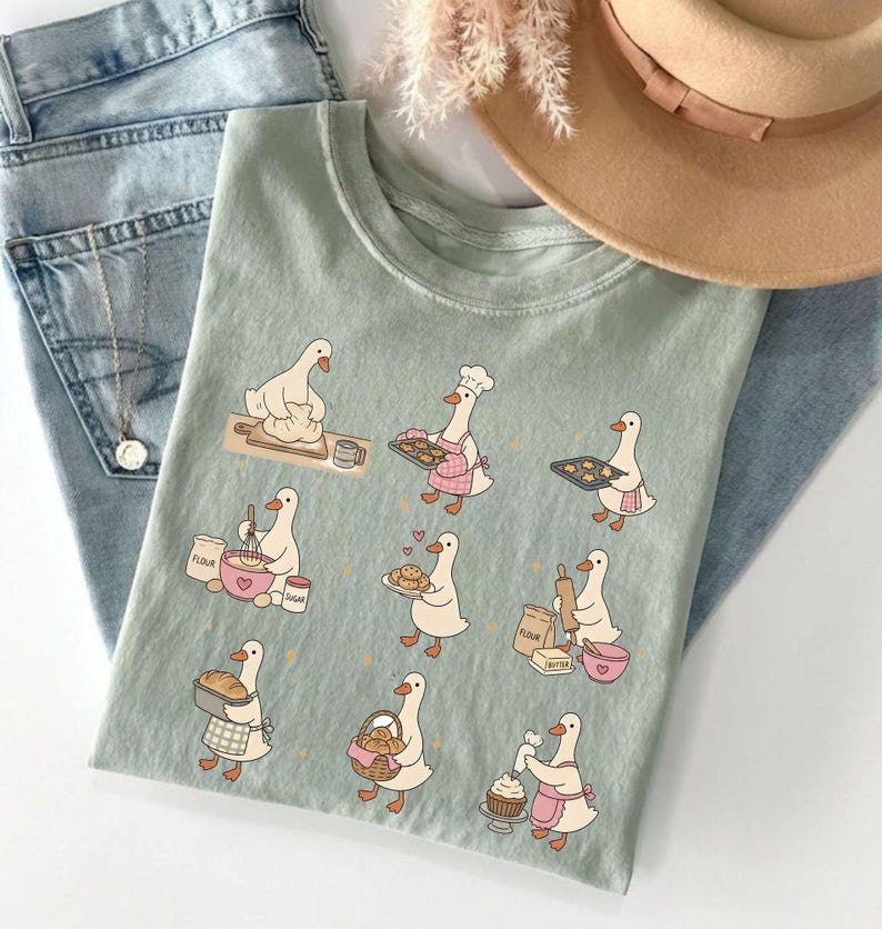 May include: A sage green t-shirt featuring a graphic print of nine white geese engaged in various baking activities, including kneading dough, mixing ingredients, holding trays of cookies, and decorating a cupcake, all in a cartoon illustration style.