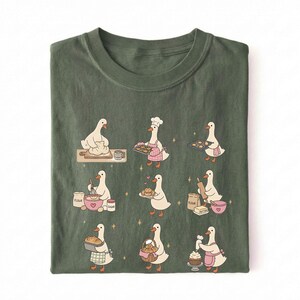 May include: An olive green t-shirt featuring a graphic print of nine white geese engaged in various baking activities, including kneading dough, wearing a chef hat, holding baking trays, whisking ingredients, and decorating a cupcake.