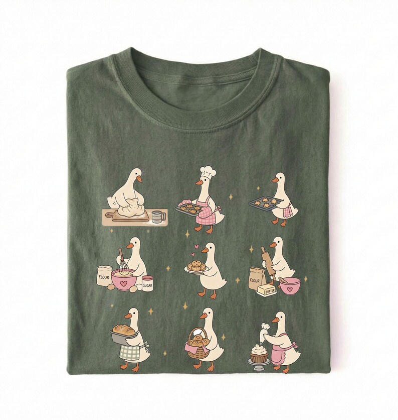 May include: An olive green t-shirt featuring a graphic print of nine white geese engaged in various baking activities, including kneading dough, wearing a chef hat, holding baking trays, whisking ingredients, and decorating a cupcake.