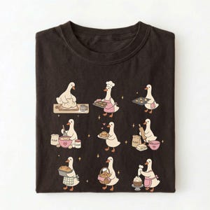 May include: A dark brown t-shirt featuring a graphic print of nine white geese engaged in various baking activities, including kneading dough, wearing a chef hat, holding trays of cookies, whisking ingredients, and decorating a cupcake.