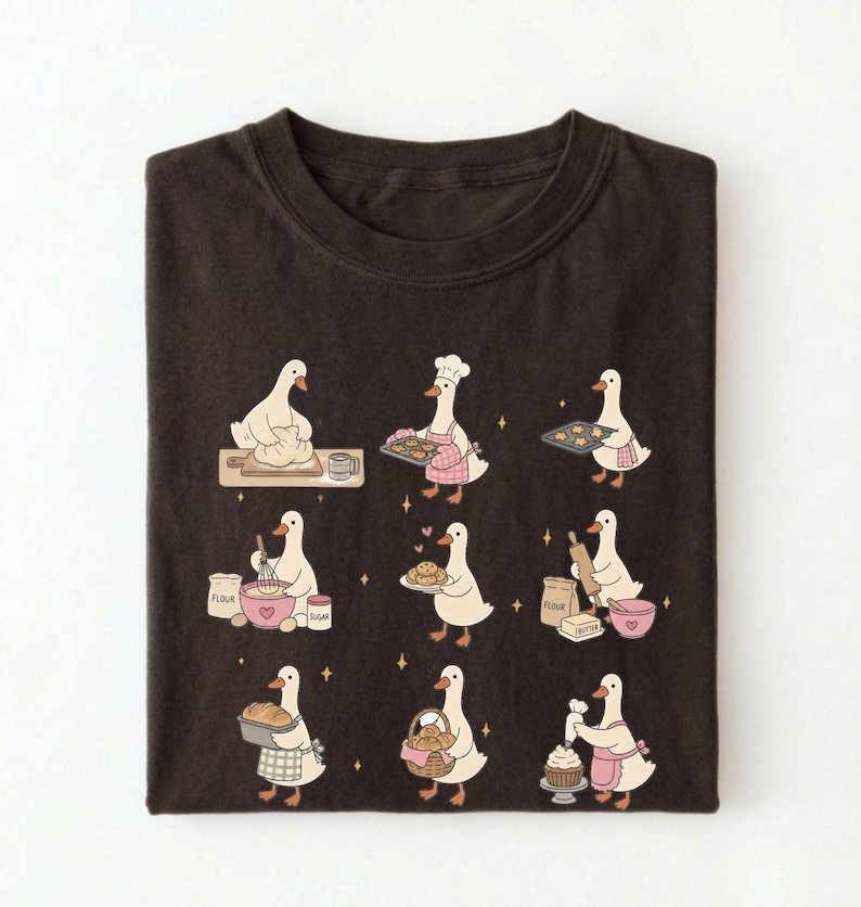 May include: A dark brown t-shirt featuring a graphic print of nine white geese engaged in various baking activities, including kneading dough, wearing a chef hat, holding trays of cookies, whisking ingredients, and decorating a cupcake.