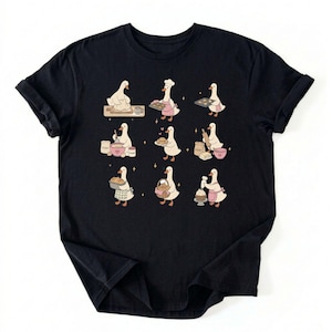 May include: A black short-sleeved t-shirt featuring a graphic print of nine white geese engaged in various baking activities. The geese are shown kneading dough, wearing chef hats, holding trays of cookies, mixing ingredients, and decorating cupcakes.