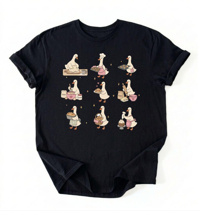 May include: A black short-sleeved t-shirt featuring a graphic print of nine white geese engaged in various baking activities. The geese are shown kneading dough, wearing chef hats, holding trays of cookies, mixing ingredients, and decorating cupcakes.