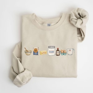 May include: A beige crewneck sweatshirt featuring a graphic print of baking ingredients in a row, including a mixing bowl with a spoon, a bag of Gold Medal flour, a stick of butter, a jar labeled SUGAR, vanilla extract, eggs, and a Pyrex measuring cup.