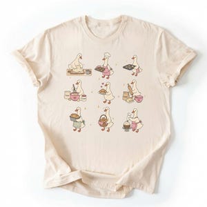 May include: A cream-colored t-shirt featuring a graphic print of nine white ducks engaged in various baking activities, including kneading dough, whisking ingredients, holding trays of cookies, rolling dough, and decorating a cupcake with pink frosting.