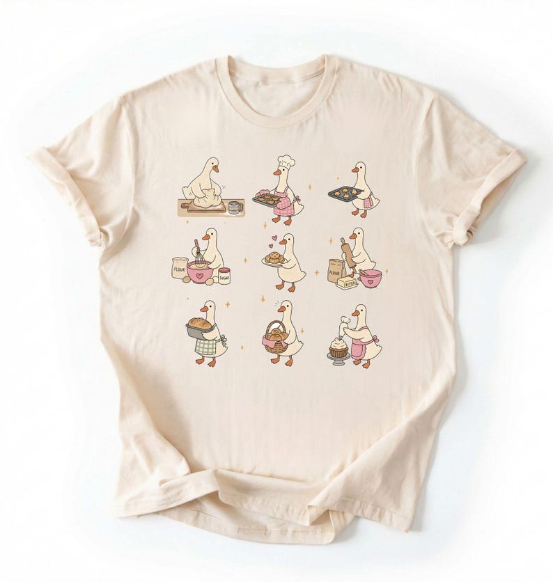 May include: A cream-colored t-shirt featuring a graphic print of nine white ducks engaged in various baking activities, including kneading dough, whisking ingredients, holding trays of cookies, rolling dough, and decorating a cupcake with pink frosting.