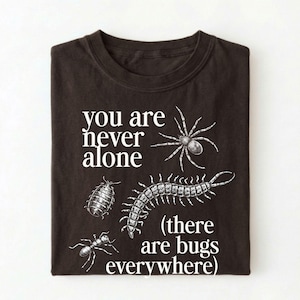 May include: A dark brown t-shirt featuring a white graphic print of a spider, a centipede, an ant, and a pill bug. The text reads, "you are never alone (there are bugs everywhere)" in a white serif font, arranged around the insect illustrations.