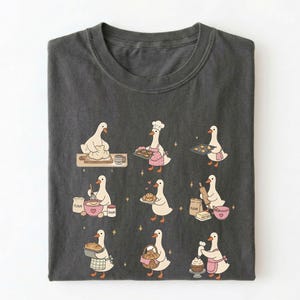 May include: A charcoal gray t-shirt featuring a graphic print of nine white geese engaged in various baking activities, including kneading dough, wearing a chef hat, holding trays of cookies, mixing ingredients, and decorating a cupcake.