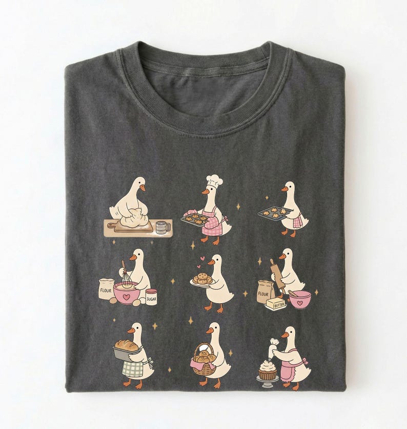 May include: A charcoal gray t-shirt featuring a graphic print of nine white geese engaged in various baking activities, including kneading dough, wearing a chef hat, holding trays of cookies, mixing ingredients, and decorating a cupcake.