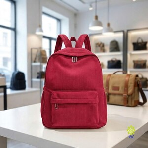 May include: A solid deep red corduroy backpack featuring a large main compartment with a dual zip closure and a smaller front pocket with a single zip. The backpack has two adjustable shoulder straps and a top handle for carrying.