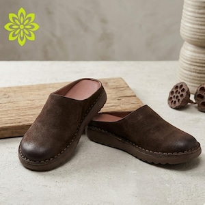 Könnte beinhalten: Ein Paar dunkelbraune Slipper aus Wildleder mit abgerundeter Zehenpartie, einer sichtbaren Naht entlang der Zwischensohle und einer flachen braunen Gummilaufsohle. Das Innenfutter der Schuhe ist in einem hellen Rosaton gehalten.