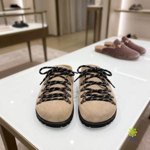 Puede incluir: Un par de zapatos sin cordones de ante beige con cordones estampados en blanco y negro pasados por ojales de metal plateado, con una suela gruesa de goma negra y un diseño de punta redondeada, expuestos sobre una superficie blanca.