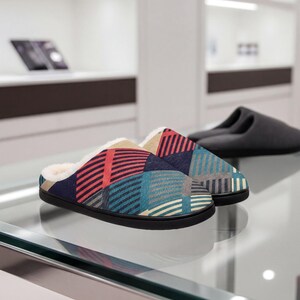 May include: A pair of slip-on slippers featuring a geometric pattern of red, blue, and beige diagonal stripes on a dark fabric upper. The interior is lined with a soft, cream-colored faux fur material, and the slippers have a solid black rubber sole.