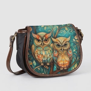 May include: A saddle-style crossbody bag featuring a front flap with a colourful illustration of two owls perched on a branch. The bag has dark brown leather-like trim, a black side panel, and an adjustable shoulder strap with gold-tone hardware.