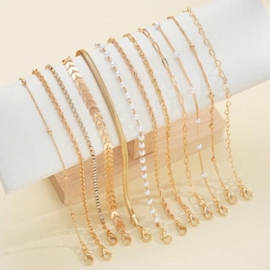 Peut inclure: Un ensemble de dix bracelets dorés présentant des designs variés, incluant une chaîne délicate avec de petites perles, un brin incrusté de cristaux, une chaîne à maillons en forme de feuille, une chaîne plate, une chaîne perlée et une chaîne à cœurs.