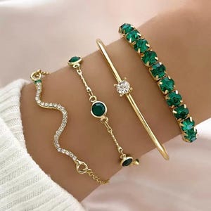 May include: A set of four gold-toned bracelets. One features a wavy snake-like design with clear crystals, another is a delicate chain with two green stones, one is a thin bangle with a single clear crystal, and the last is a row of green crystals.