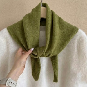 May include: A solid olive green knit scarf draped over a white fuzzy sweater. The scarf features a soft, textured fabric with a triangular shape, tied in a simple knot at the center, hanging down in two distinct ends against the white garment.
