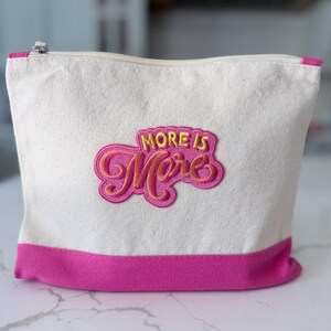 May include: A cream-colored canvas zippered pouch featuring a bright pink bottom border and a central embroidered patch. The patch displays the text "MORE IS More" in yellow and orange cursive lettering against a vibrant pink background.