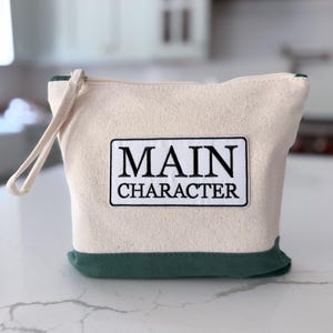 Puede incluir: Un estuche de lona color crema con cremallera, base verde oscuro y una correa de muñeca a juego. En el centro de la parte frontal hay un parche rectangular blanco con el texto negro MAIN CHARACTER en una fuente serif negrita y borde fino.