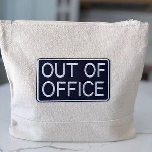 Può includere: Una pochette in tela color naturale con chiusura a zip e una toppa rettangolare nera centrata sul davanti. La toppa riporta la scritta ricamata in bianco OUT OF OFFICE in un carattere sans-serif pulito, creando un netto contrasto col tessuto.