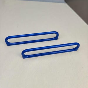 Puede incluir: Dos tiradores de armario alargados de color azul brillante con un acabado liso y lustroso. Cada tirador presenta un diseño de bucle rectangular redondeado continuo con un centro hueco, proporcionando un aspecto elegante y uniforme.