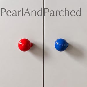 Pode incluir: Dois puxadores de armário esféricos fixados em portas de armário brancas. Um puxador é vermelho brilhante e o outro é azul brilhante. O texto PearlAndParched é visível em letras cinzentas no topo das portas de armário brancas.
