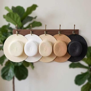 May include: A horizontal wooden wall rack with five brass-colored metal hooks. Hanging from the hooks are five circular straw hats in a gradient of colors, ranging from cream and white to light tan, medium brown, and solid black.