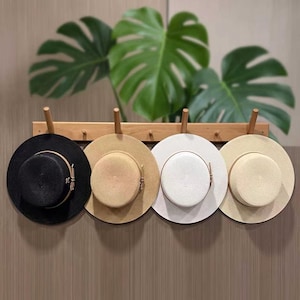 May include: Four straw hats hanging on a wooden wall rack. The hats are black, tan, white, and light beige, each featuring a flat crown, a wide brim, and a thin decorative band around the base of the crown. The rack has four wooden pegs.