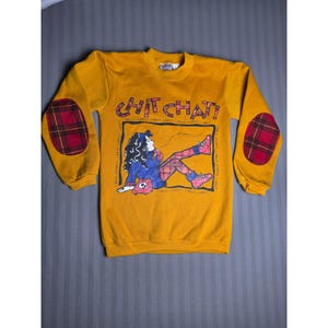 May include: A mustard yellow long-sleeve sweatshirt featuring red and black plaid elbow patches. The front displays a graphic of a person with dark hair on a telephone with the text CHIT CHAT! and repeated JABBER text in a square frame.